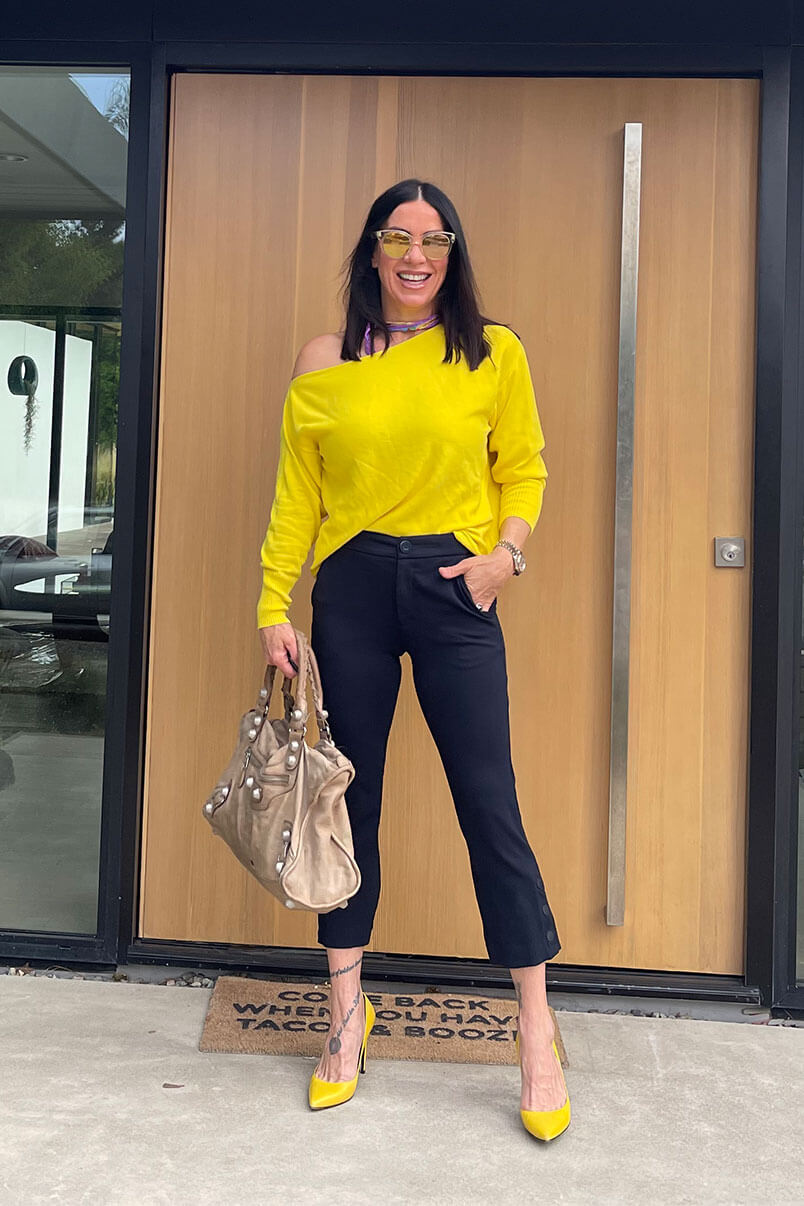 Kat Woodside, Chief Design Officer models her outfit of the day featuring the Buttercup Pullover in Neon Yellow, Black Tie Trouser in Black, statement accessories and heels.